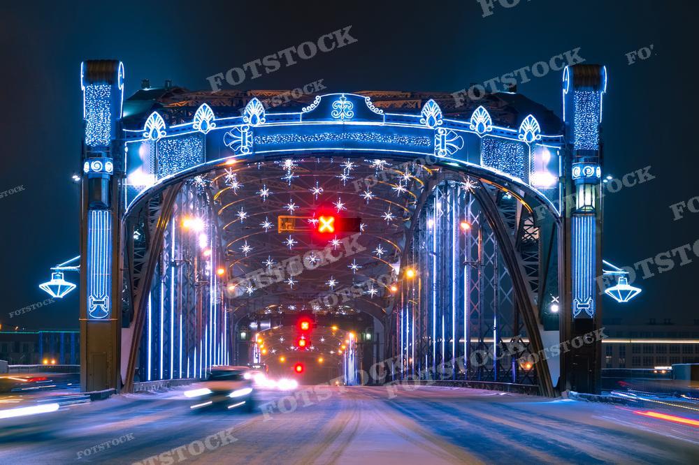 На фотографии — заснеженный мост в Санкт-Петербурге, украшенный новогодней иллюминацией. Яркие огни создают праздничное настроение и красиво контрастируют с темным небом и снегом. Это изображение может использоваться для новогодних поздравлений, рекламных кампаний зимних праздников или в туристических проспектах, показывающих красоту города в зимнее время.